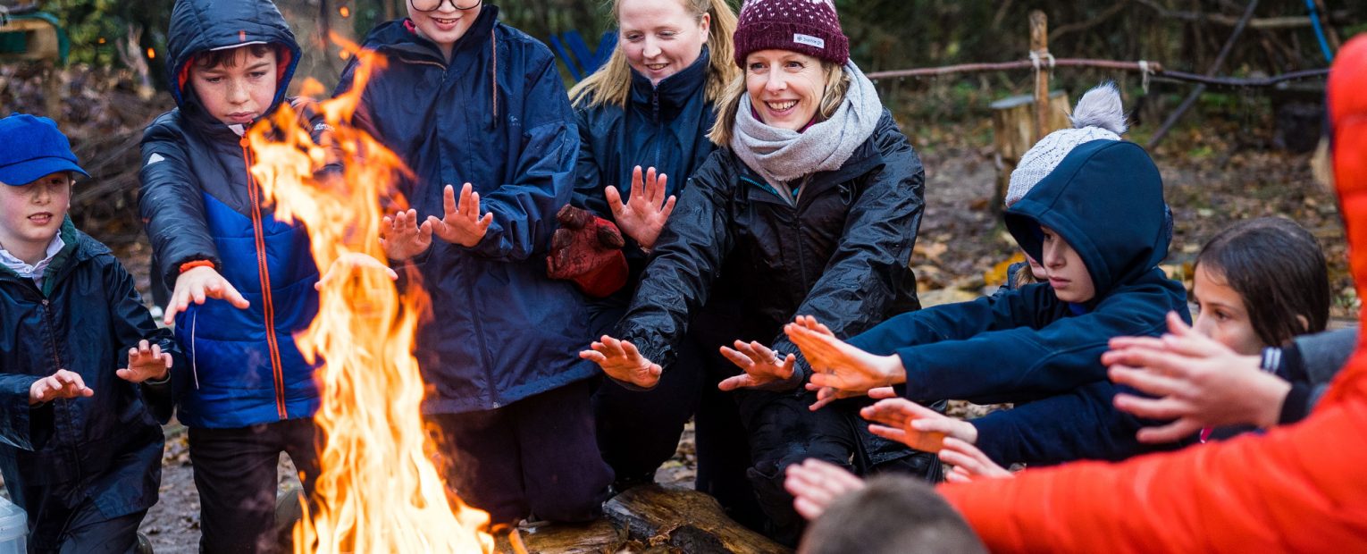 A teacher and their students holding their hands towards a campfire to keep warm